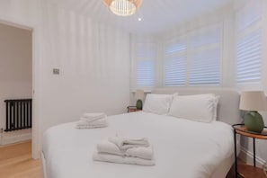 A hotel room with a large bed, a bedside table, a lamp, and a window with blinds. - Stylish Garden Home 2 BDR Wimbledon (London)