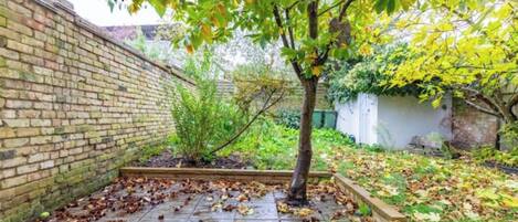 Un mur de briques, un arbre et un espace jardin parsemé de feuilles mortes.