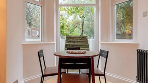 A dining area with a table and chairs, a view of a garden through a window, and a radiator on the wall. - Stylish Garden Home 2 BDR Wimbledon (London)