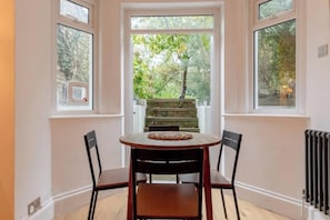 A dining area with a table and chairs, a view of a garden through a window, and a radiator on the wall. - Stylish Garden Home 2 BDR Wimbledon (London)
