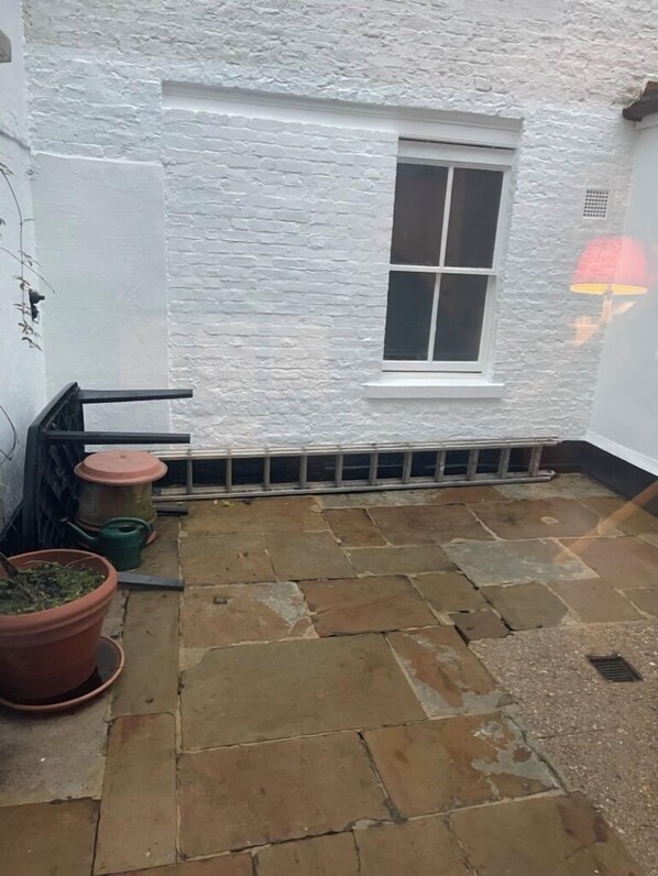 A small, white-walled room with a single window, a potted plant, and a stone floor.