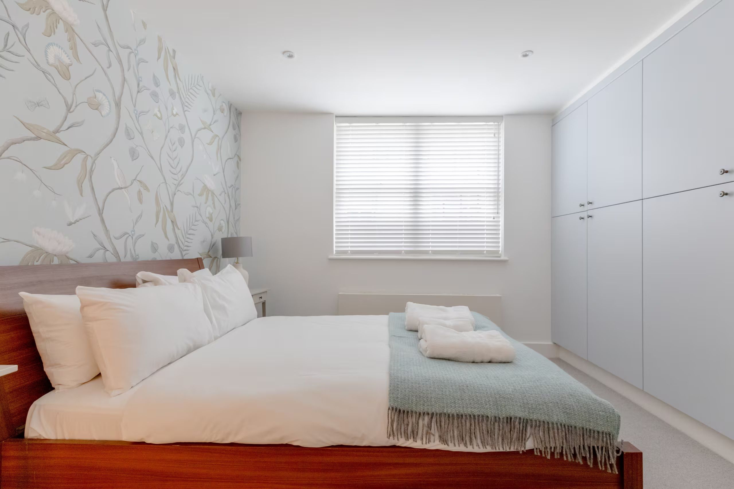 Une chambre à coucher comprenant un lit, une table de chevet, une lampe, une fenêtre avec des stores et une armoire.