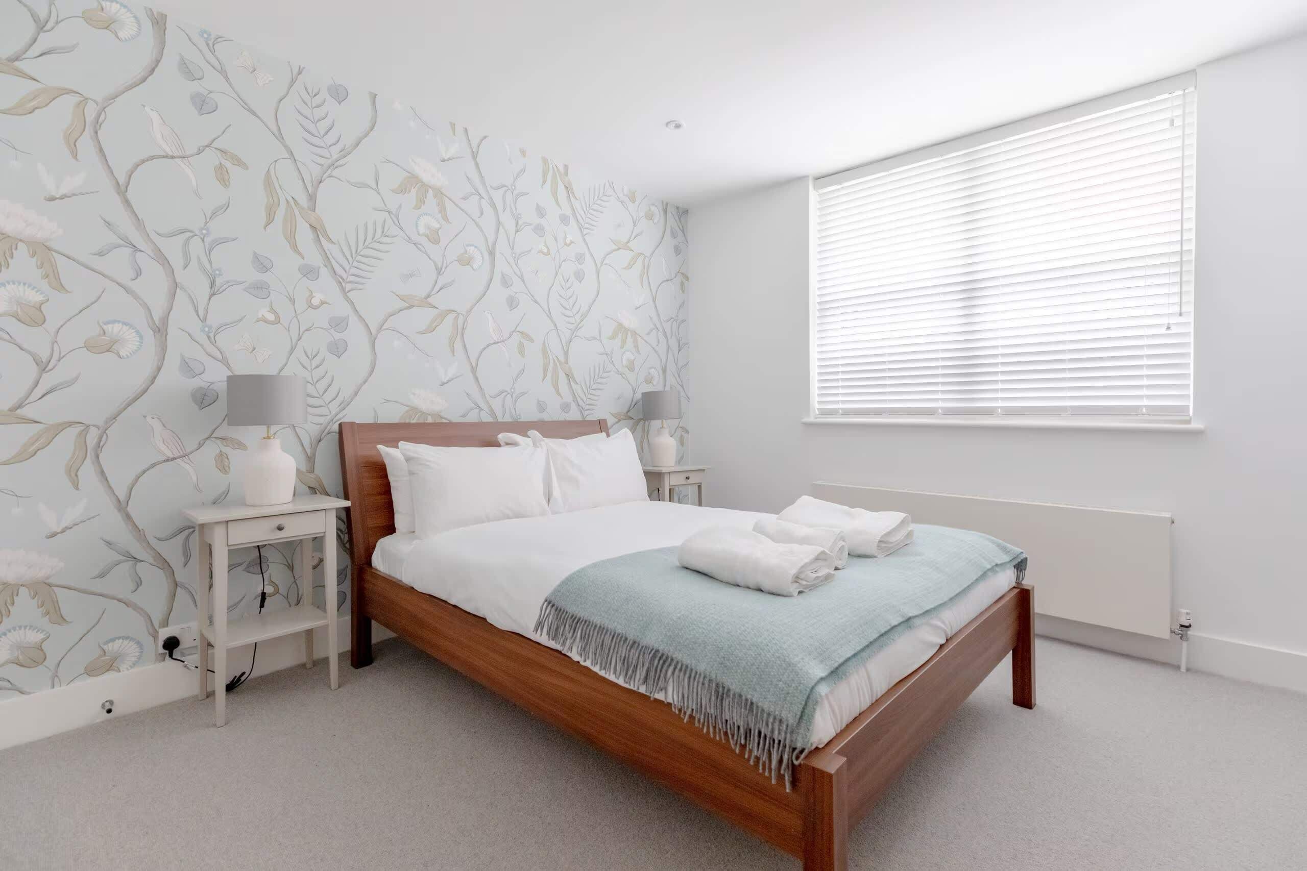 Un dormitorio con cama de madera, ropa de cama blanca, una manta azul, una mesita de noche con lámpara y una ventana con persianas.