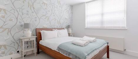Un dormitorio con cama de madera, ropa de cama blanca, una manta azul, una mesita de noche con lámpara y una ventana con persianas.