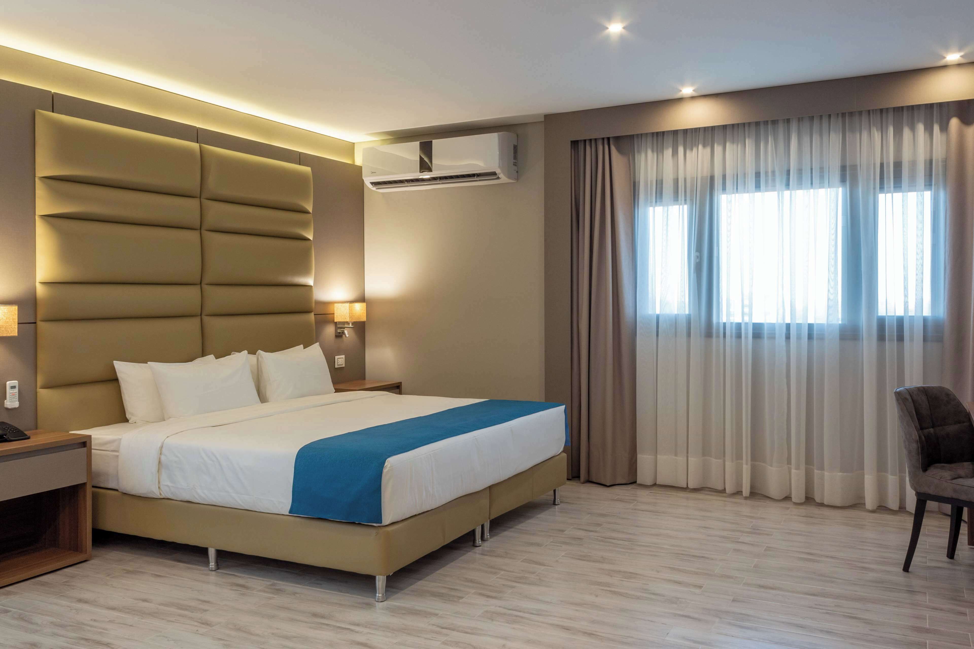 Una habitación de hotel moderna con una cama grande, un equipo de aire acondicionado, una lámpara de noche, una silla y una ventana con cortinas.