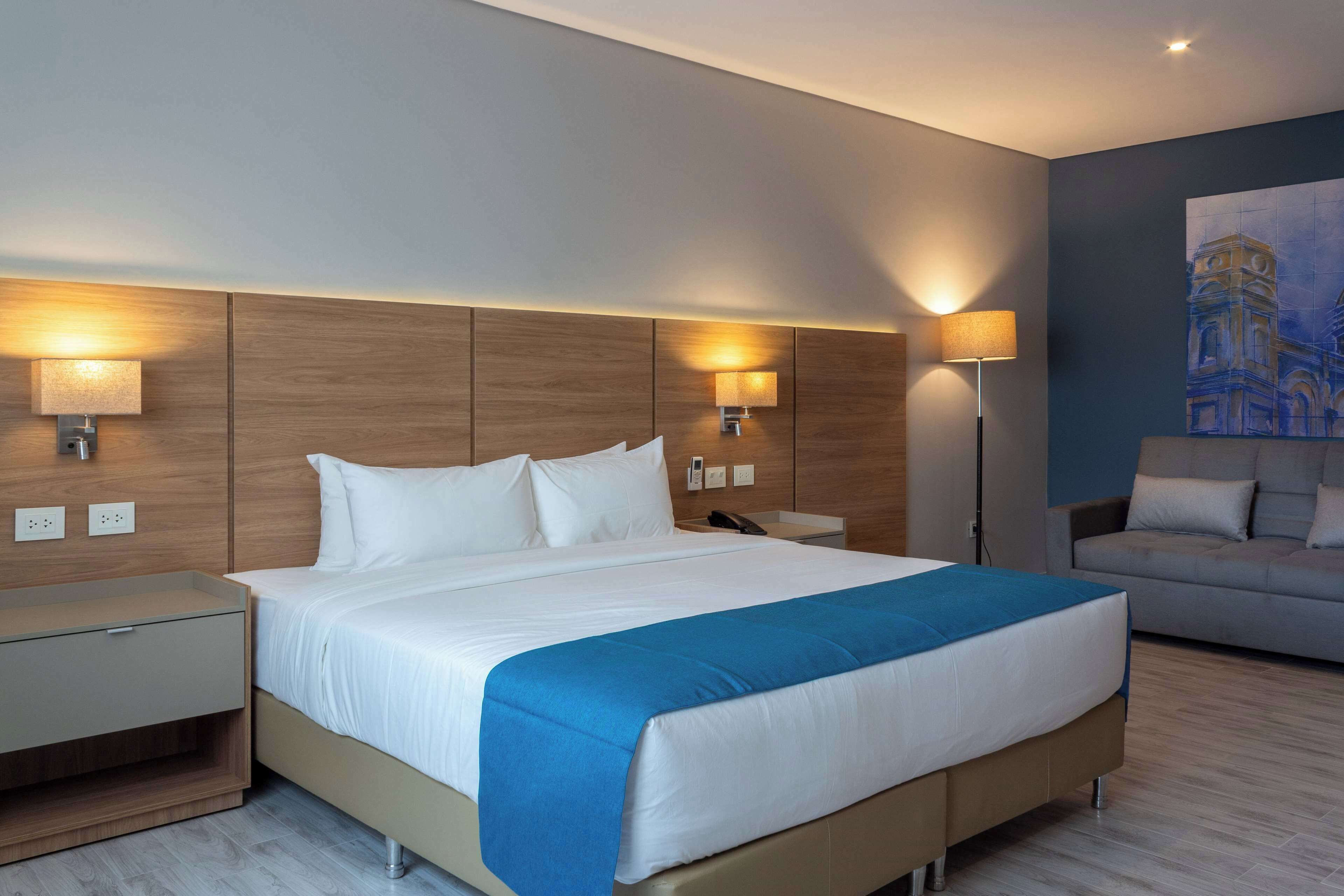 Moderni hotellihuone, jossa on suuri sänky, yöpöydällä olevat lamput, työpöytä ja sohva.