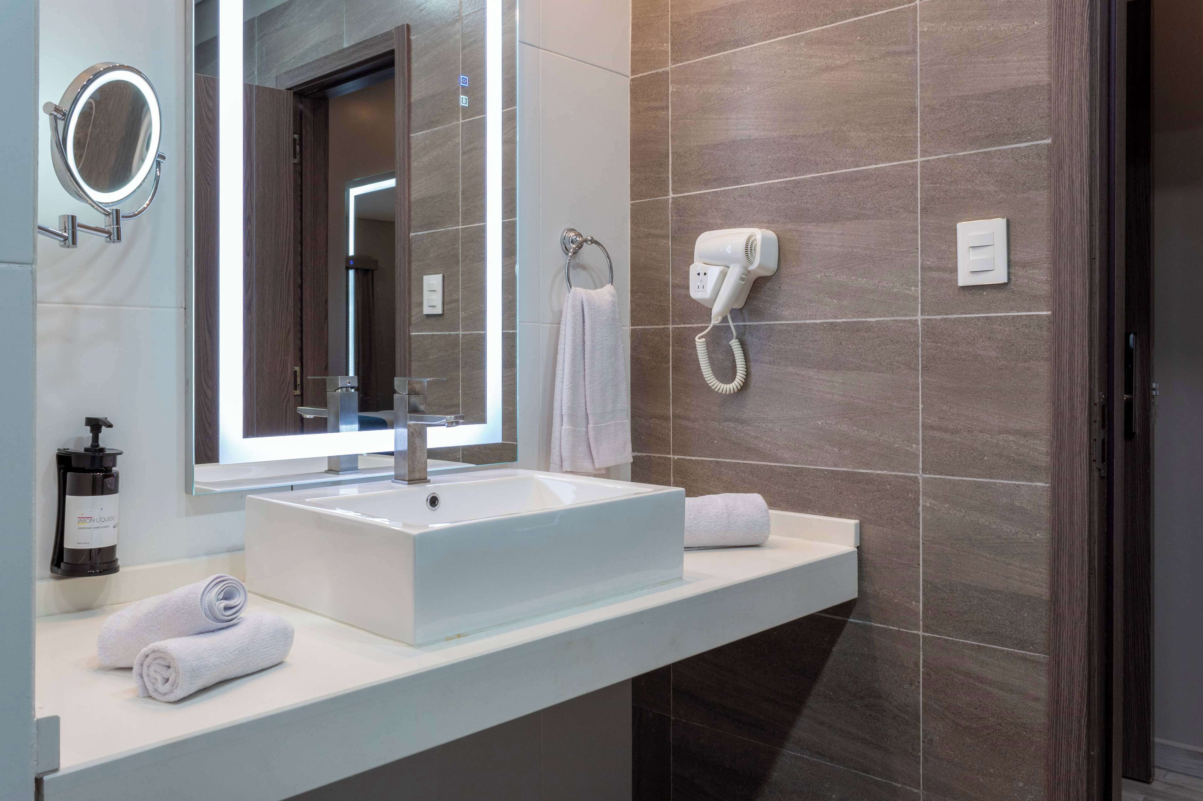 Une salle de bain moderne avec un grand miroir, un lavabo blanc, un sèche-cheveux et des serviettes soigneusement pliées sur le comptoir.