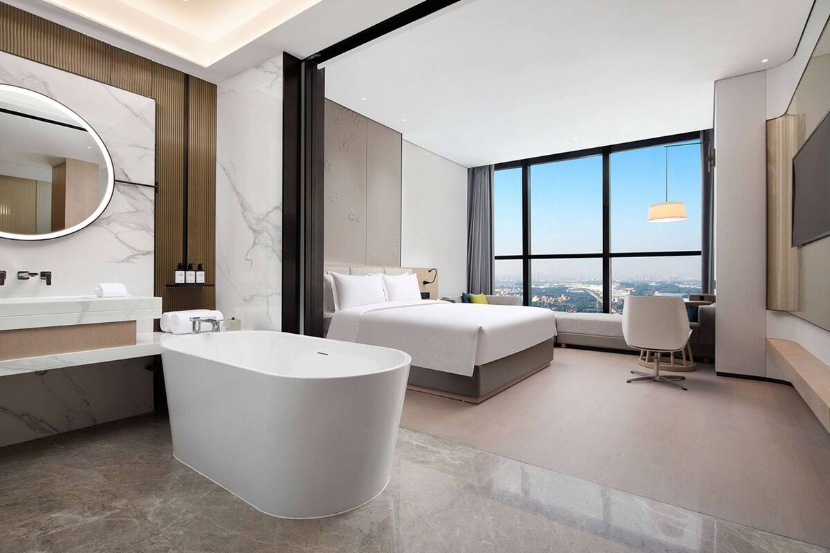 A modern hotel room with a large bathtub, a bed, a desk, and a large window offering a city view.