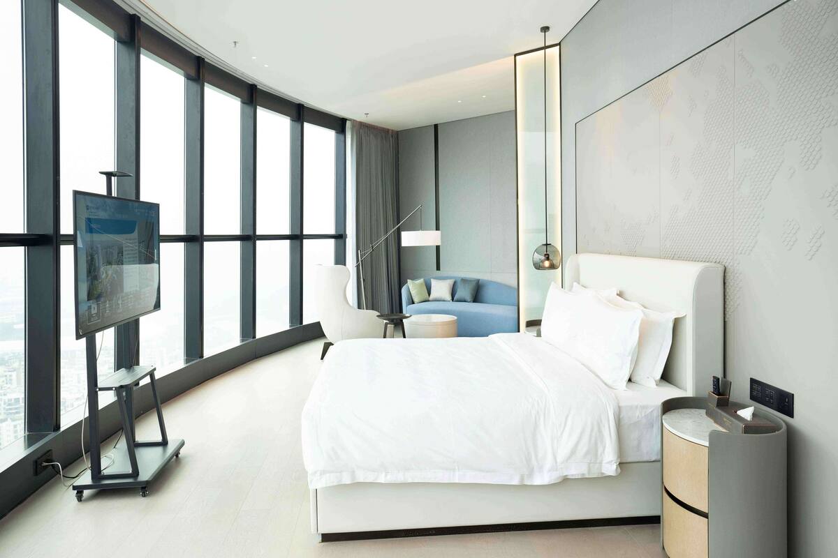 A modern hotel room with a large bed, a TV on a stand, a sofa, and a view of the city through large windows.