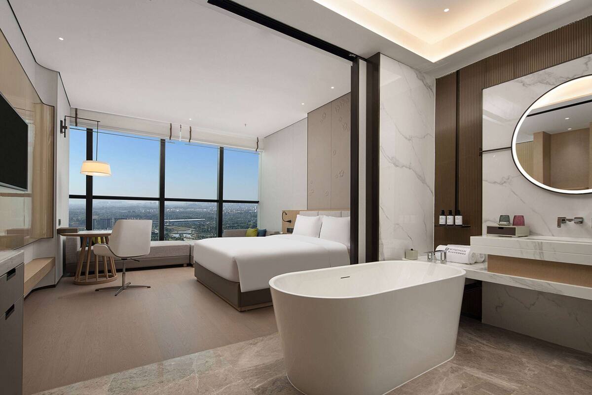 A modern hotel room with a large bed, a bathtub, a desk with a chair, a large window with a city view, and a bathroom with a mirror and sink.