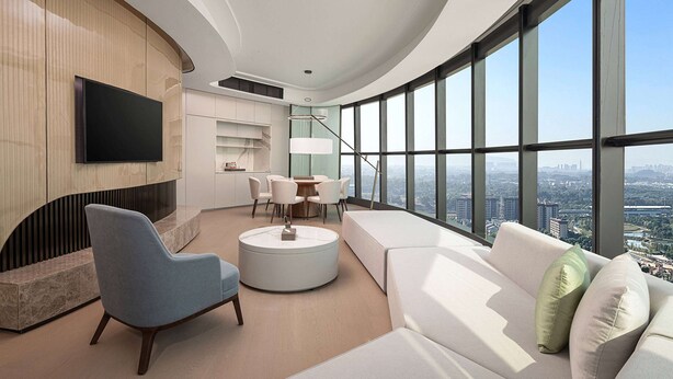 A modern living room with a curved ceiling, a flat-screen TV mounted on the wall, a seating area with a sofa and armchairs, a round coffee table, and a view of the city through large windows. - Wyndham Foshan Nanhai (Foshan)