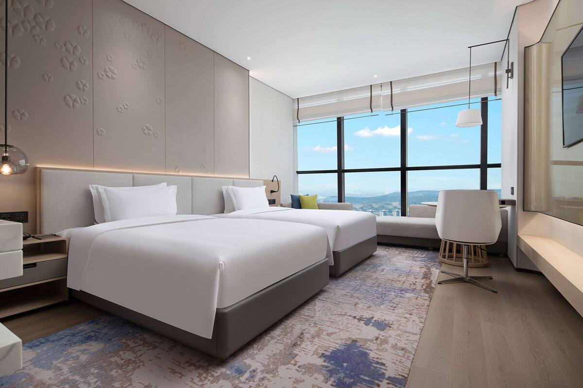 A modern hotel room with a large bed, a seating area, a desk, and a large window offering a city view.
