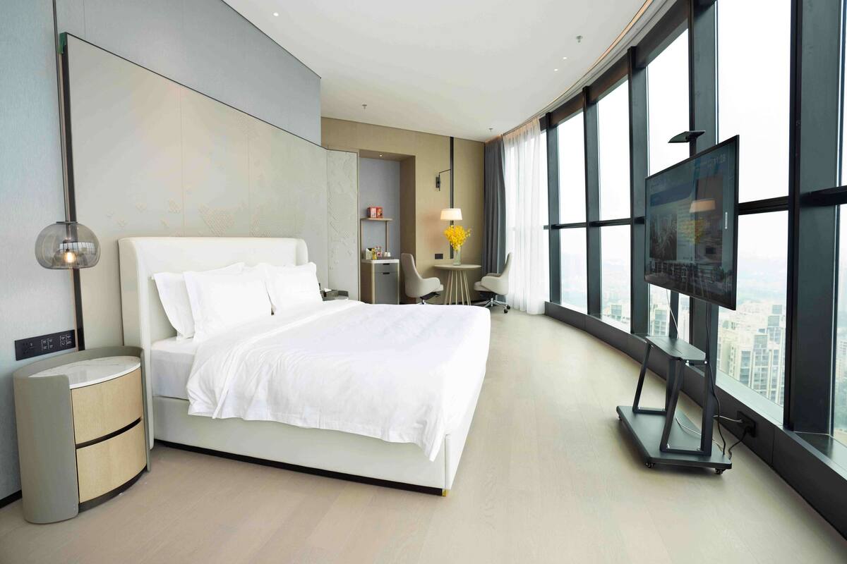 A modern hotel room with a large bed, a desk, a chair, a TV, and a view of the city through large windows.