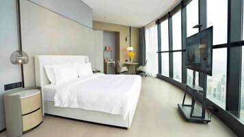 A modern hotel room with a large bed, a desk, a chair, a TV, and a view of the city through large windows.