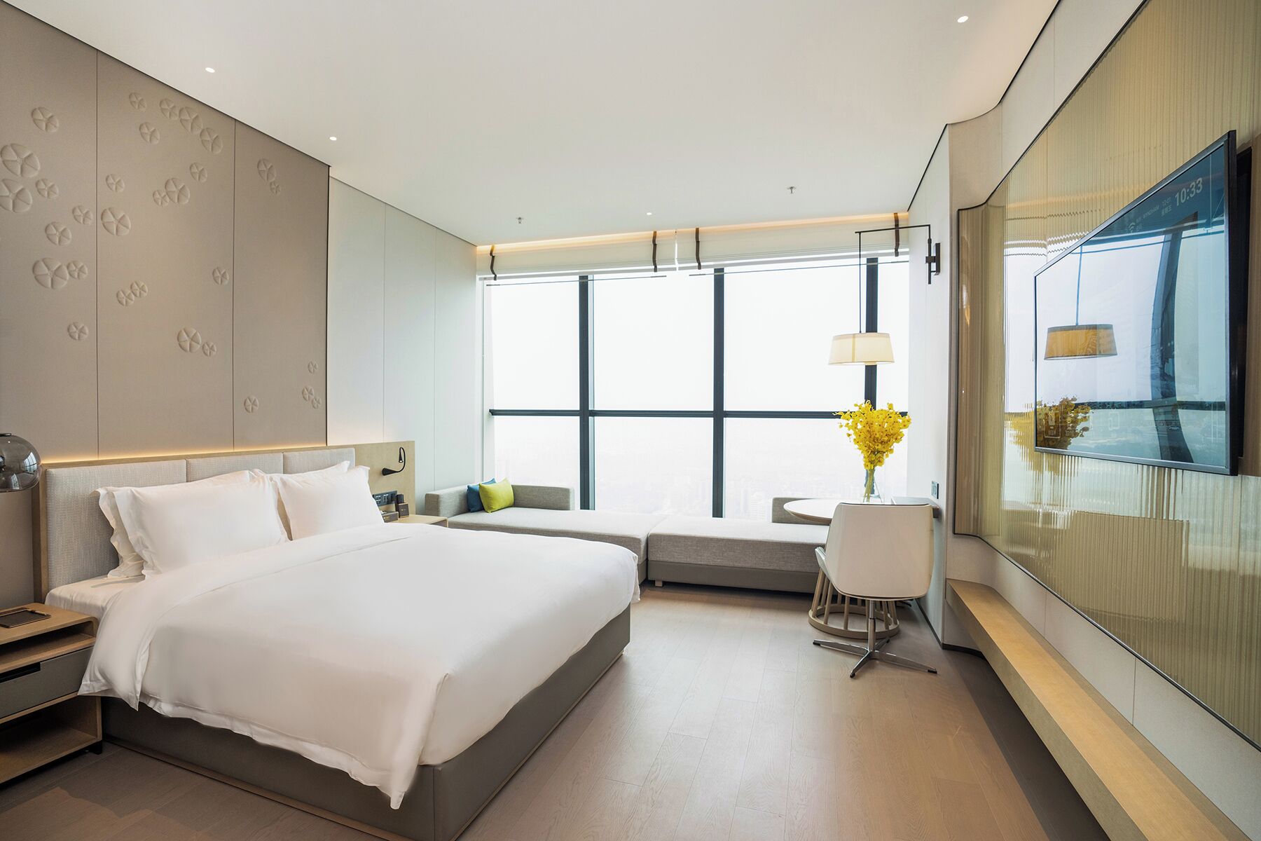 Une chambre d’hôtel moderne dotée d’un grand lit, d’un coin salon et offrant une vue sur la ville grâce à de grandes fenêtres.