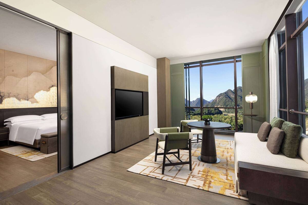 Una habitación de hotel moderna con cama, televisor, sofá y zona de comedor con sillas y mesa.