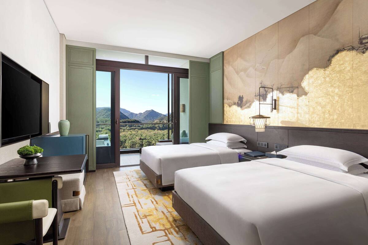 Habitación de hotel con dos camas, un escritorio, una silla y un amplio ventanal con vistas a la montaña.