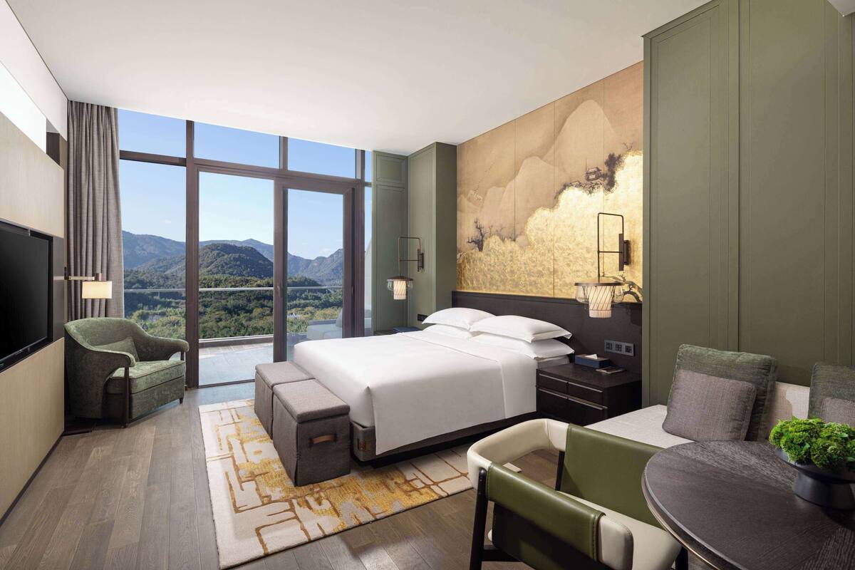 Una habitación de hotel moderna con una cama grande, un área de descanso y vistas a las montañas a través de amplias ventanas.