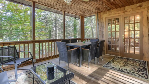 Une véranda abritée avec une table et des chaises, un ventilateur de plafond et une vue sur la forêt.