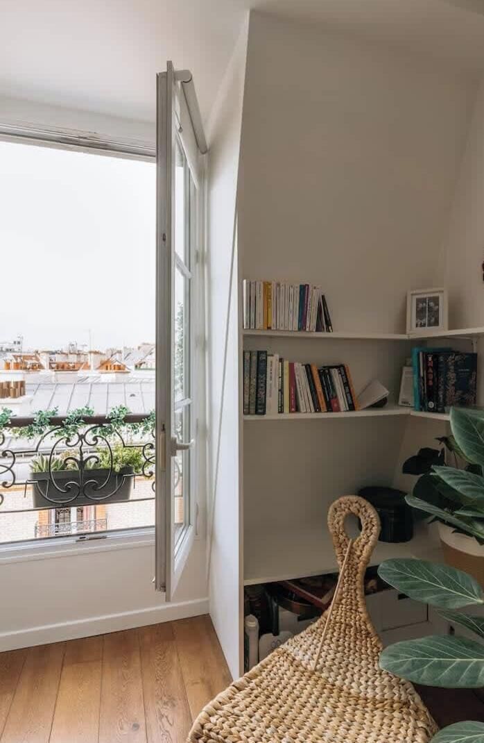 Ein Zimmer mit einem großen Fenster, einem Bücherregal und einem gewebten Stuhl.