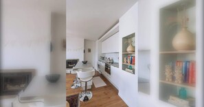 Una cocina moderna con armarios blancos, un comedor con mesa y sillas blancas, y una estantería con objetos decorativos.
