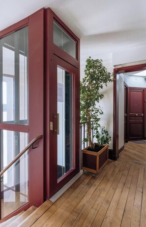 A hallway with a wooden floor, a red door with glass panels, and a potted plant. - Top Floor 2BD Apartment Amazing Views—Montmartre! (Paris)