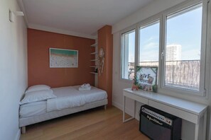A bedroom with a bed, a desk with a speaker, a window with a view, and a wall decoration. - Breathtaking 2BR – Buttes Chaumont & Terraces (Paris)