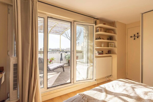 A bedroom with a sliding glass door to a balcony, a radiator, a bed with a white blanket, and a shelf with various items.