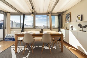 A modern room with a large table, two chairs, a hanging light fixture, and a city view through large windows.