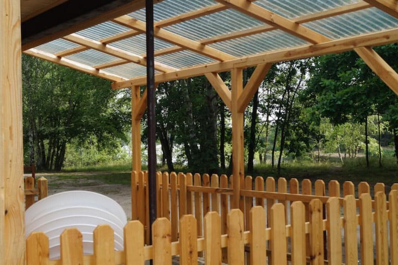 A wooden gazebo with a metal roof, surrounded by a wooden fence, set in a wooded area.