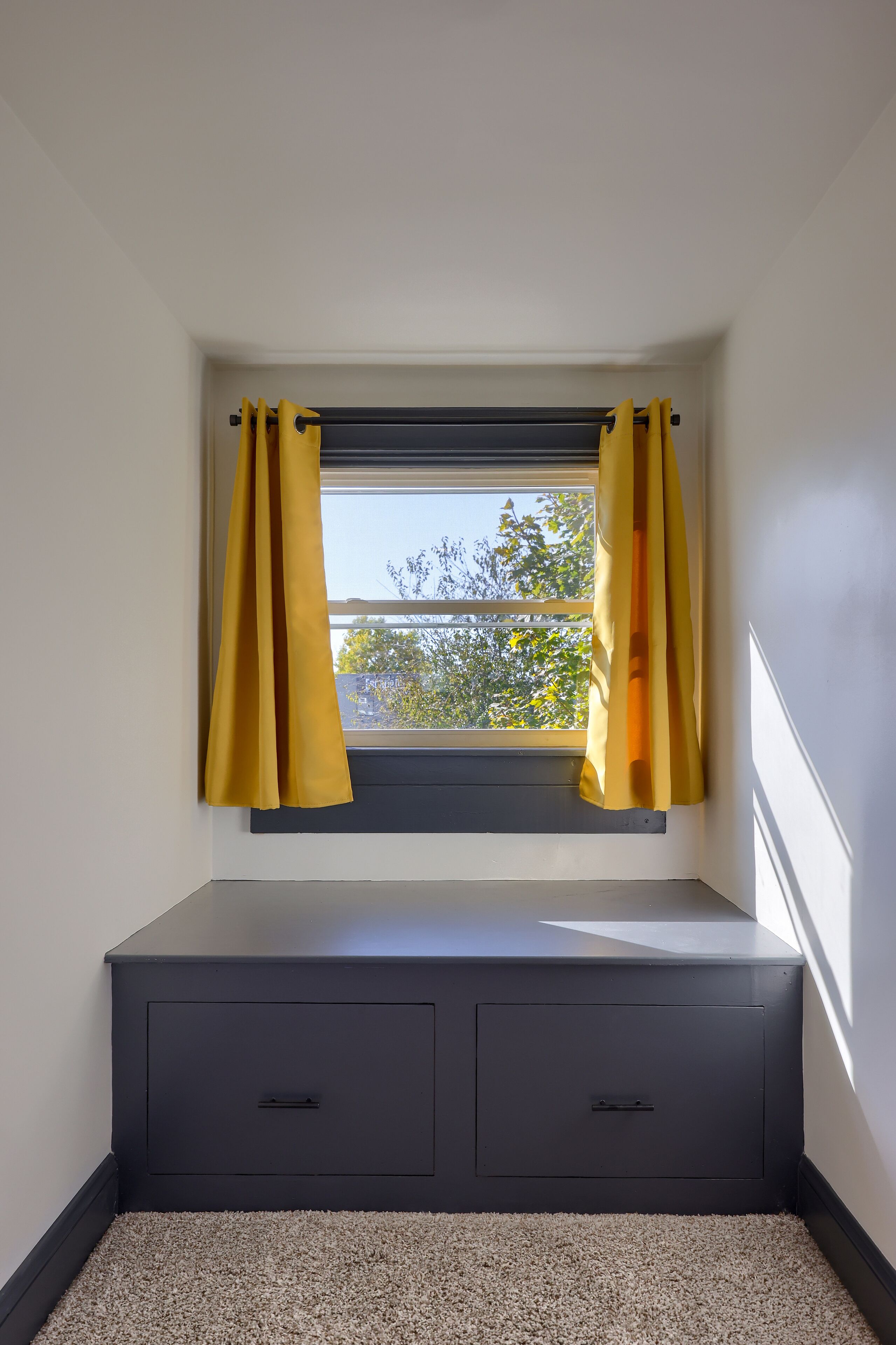 Een kamer met een raam, daaronder een donker opbergvak, en een vloerbedekking.