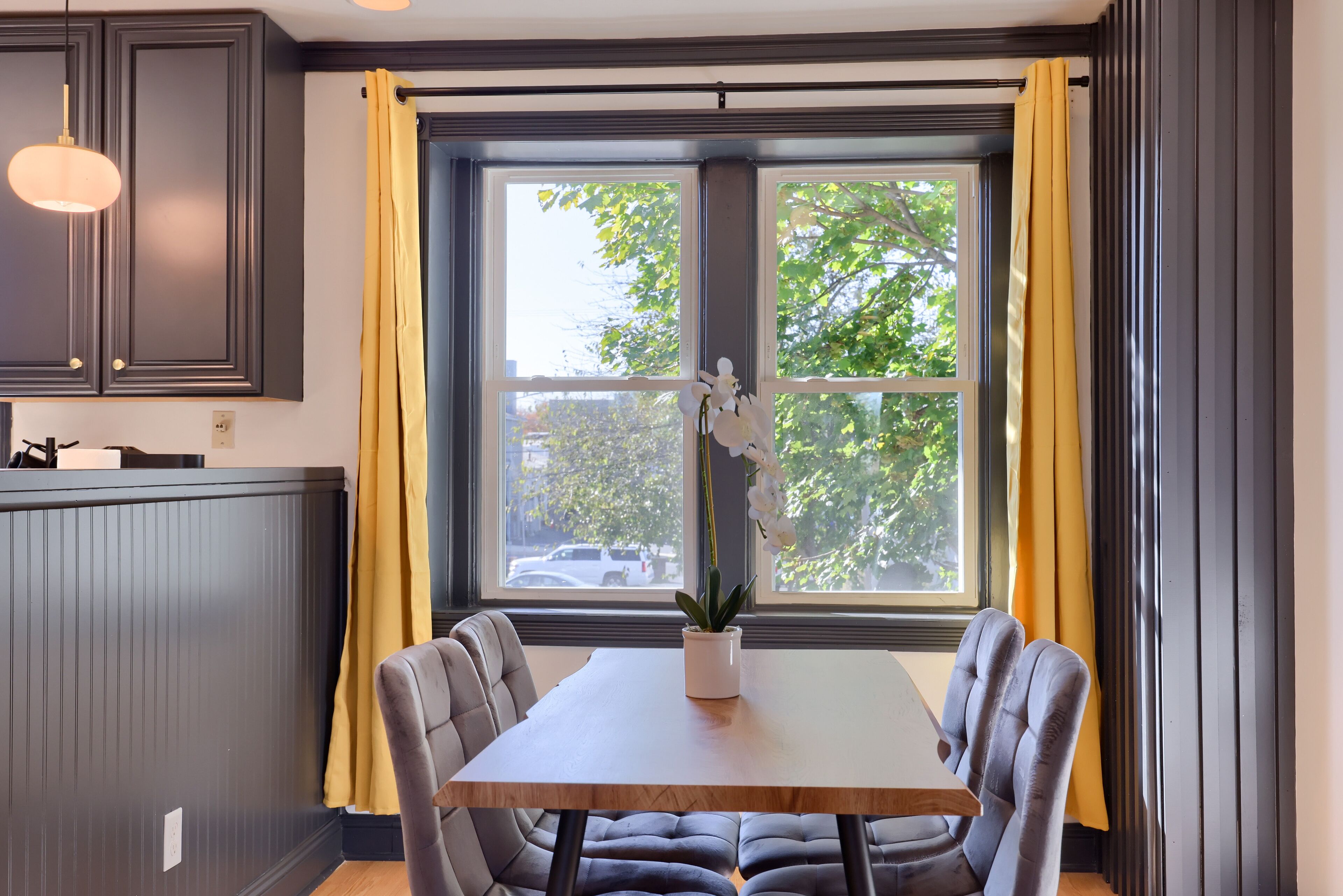 Un coin repas avec une table en bois et des chaises rembourrées, une plante en pot et une vue sur les arbres à travers une grande fenêtre ornée de rideaux jaunes.