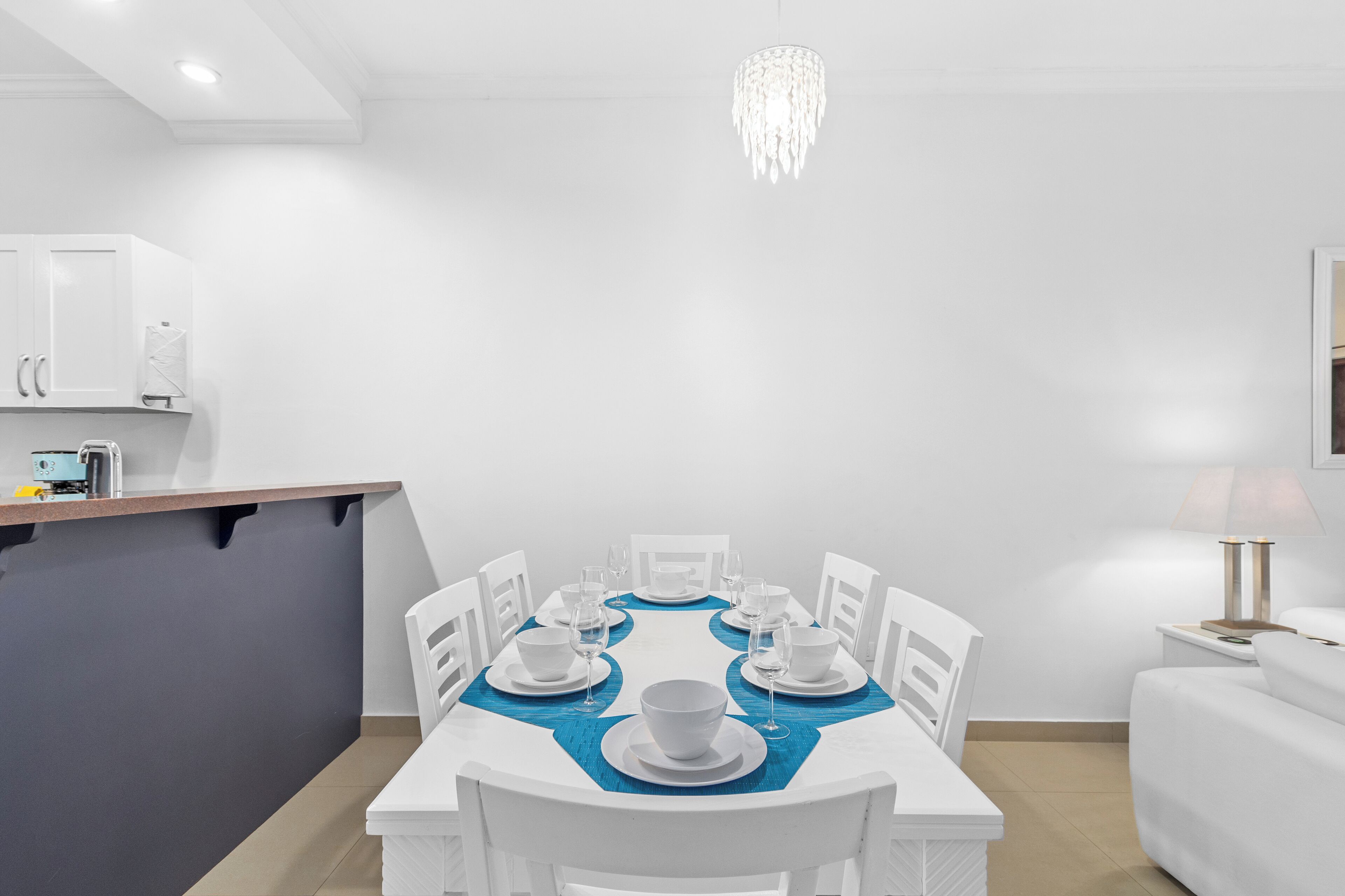 A modern, white dining area with a round table set for six, a kitchen counter, and a bedside table with a lamp.