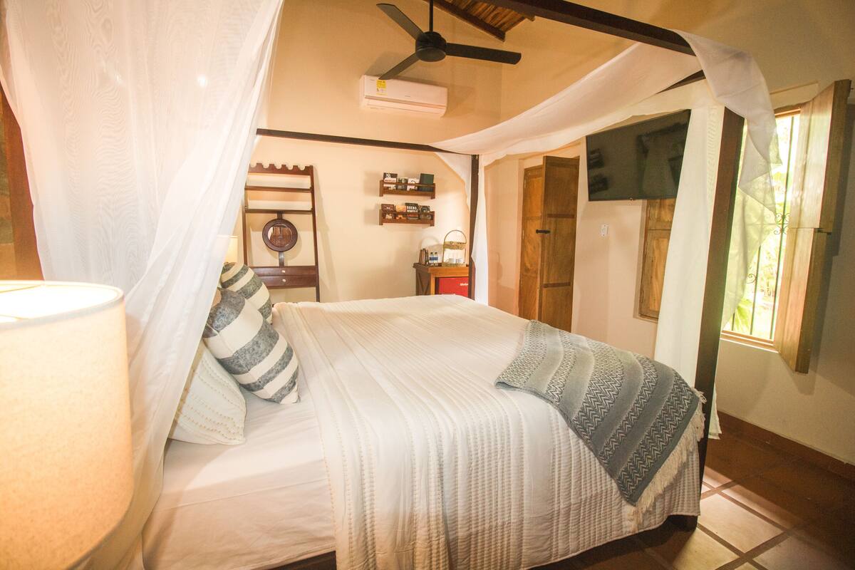 Un dormitorio con cama, ventilador de techo, aire acondicionado, mesita de noche, lámpara y ventana con cortinas.