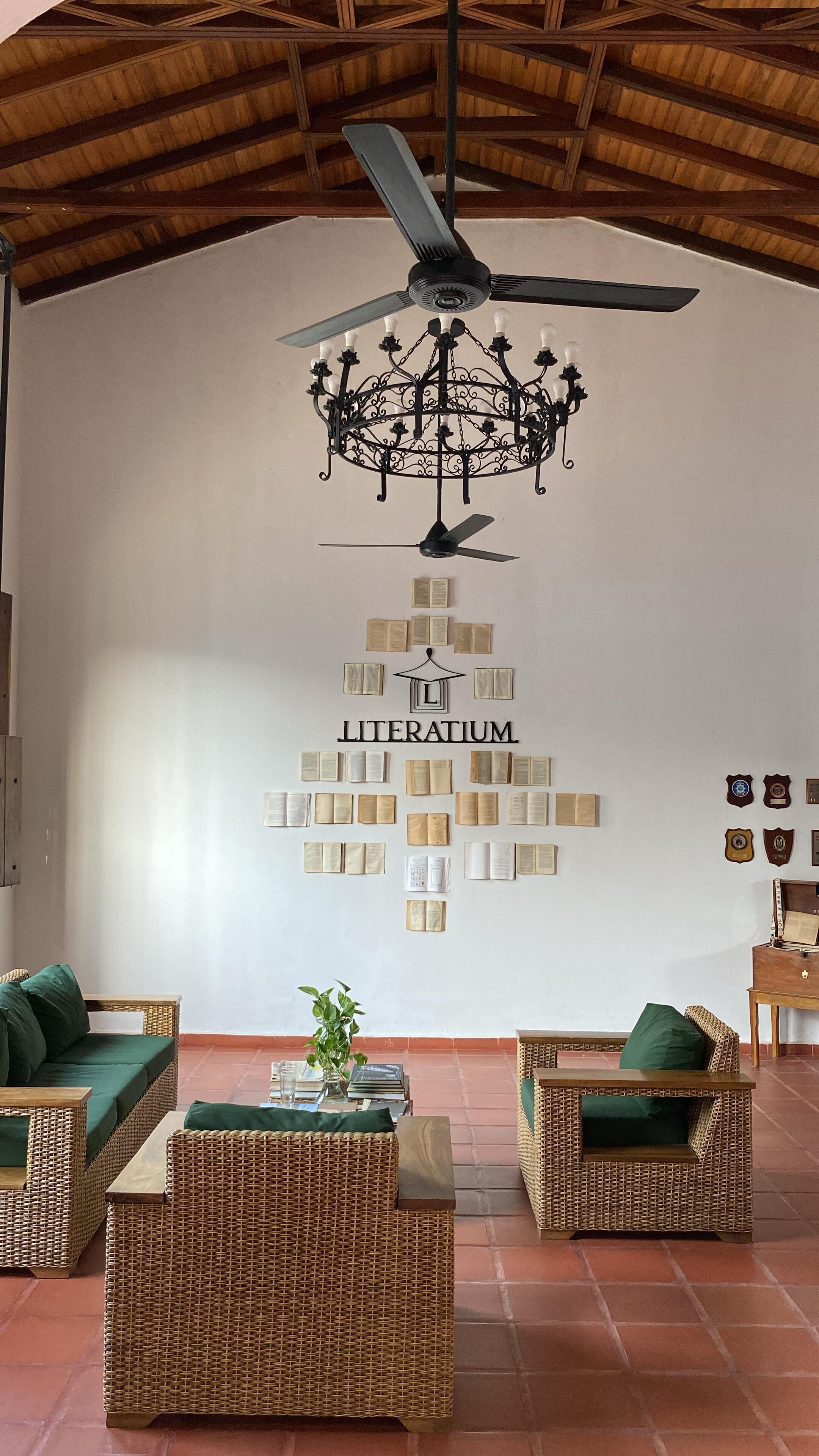 Et værelse med en loftsvifte, en lysekrone og en væg dekoreret med bøger samt et 'LITERATUM' skilt. Der er møbler af flet og et lille bord med en plante.