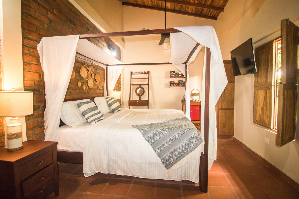 Un dormitorio con una cama con dosel, una mesita de noche, una lámpara, una pared de ladrillo, un techo de madera y un televisor montado en la pared.