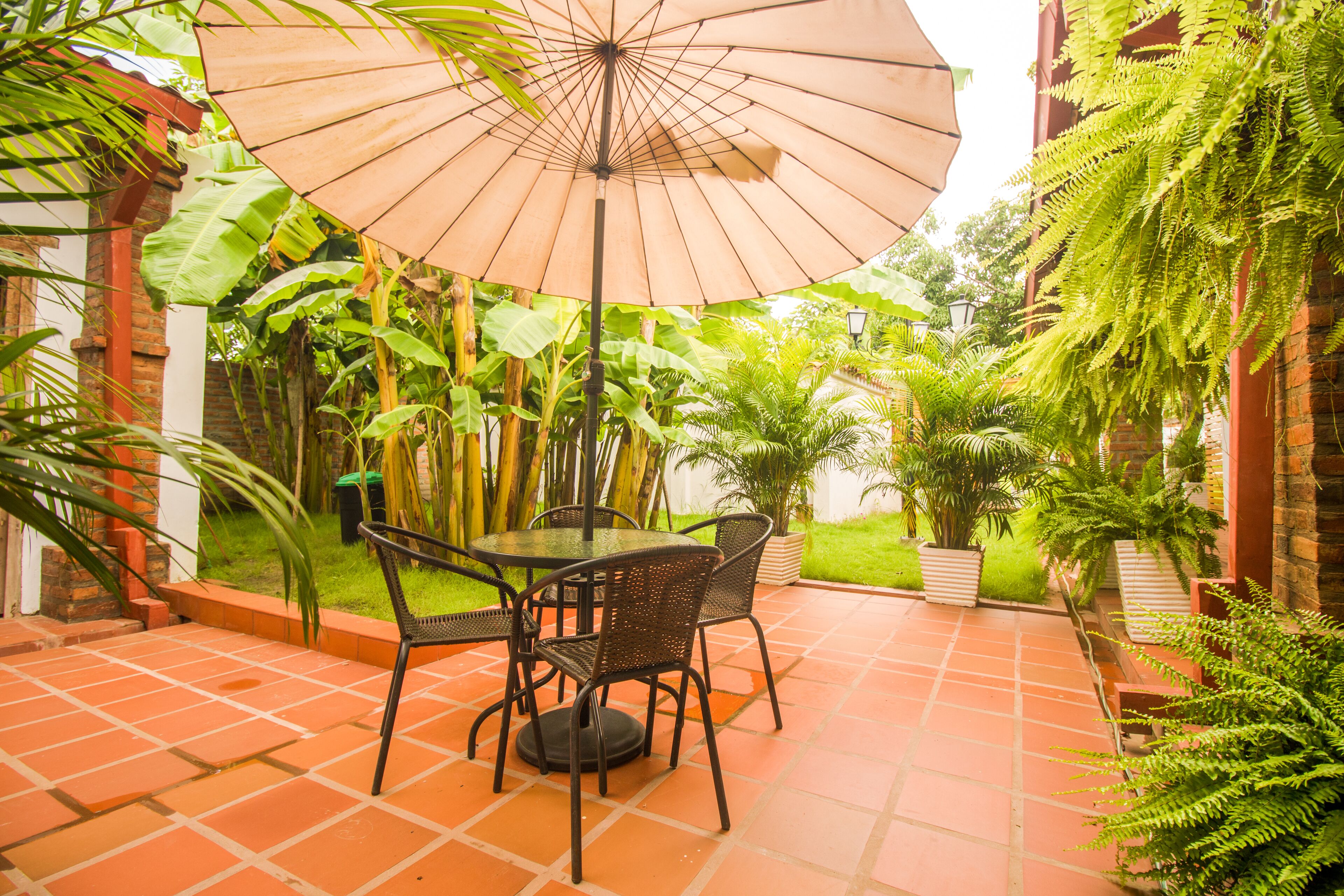A patio with a table and chairs, surrounded by lush greenery and a large umbrella.