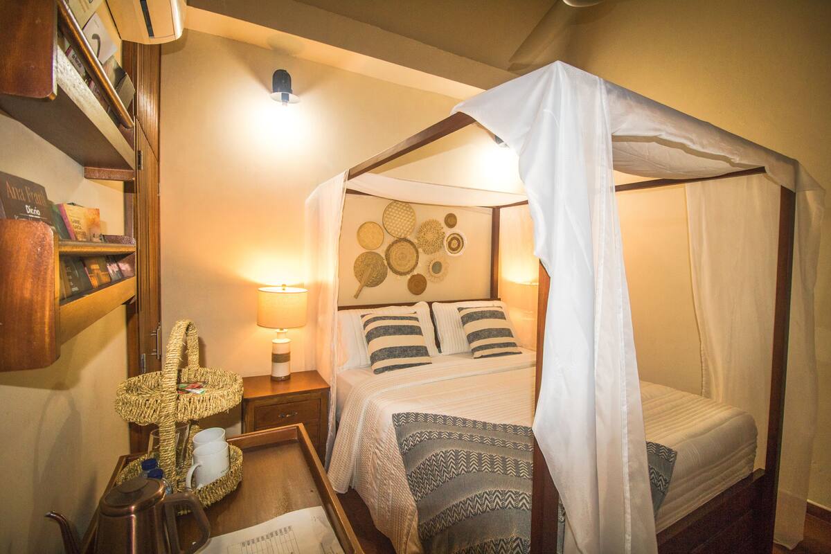 Un dormitorio con cama de dosel, una mesita de noche con lámpara, una cesta de mimbre y una estantería.