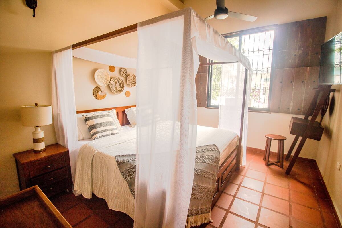 Una cama con dosel y cuatro postes, una mesita de noche con lámpara, un taburete pequeño y una pared decorada con placas.