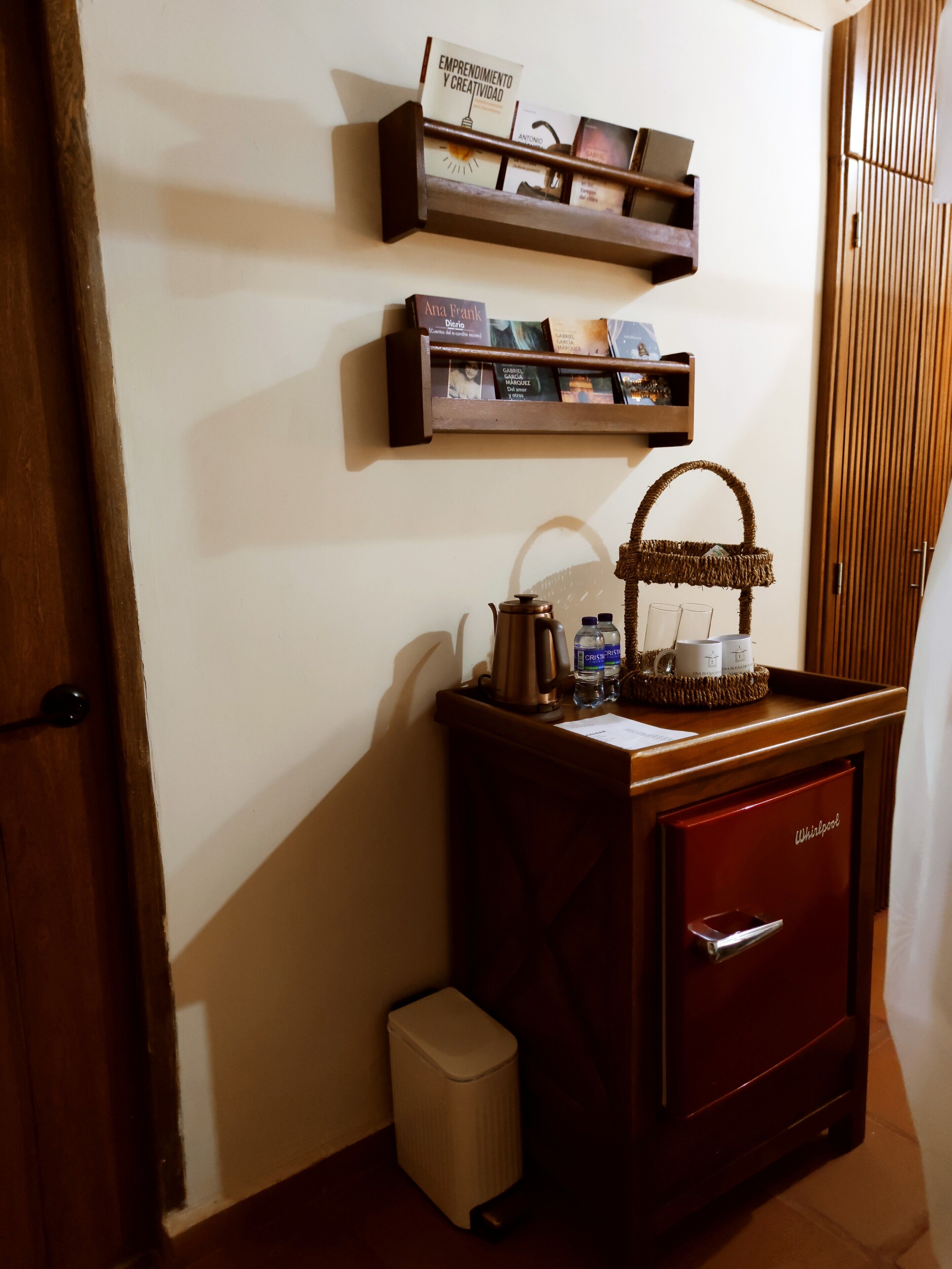 Een kitchenette met een koffiezetapparaat, waterflesjes en een kleine prullenbak. Boven het aanrecht staan houten planken met boeken en een bordje.