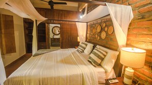 Un lit bien fait, recouvert d’une couvre-lit à rayures, une table de chevet avec une lampe, et un mur en briques agrémenté de décorations en bois.