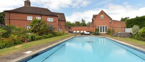 A rectangular swimming pool with a tiled edge, surrounded by a grassy area and a brick building with a pitched roof.