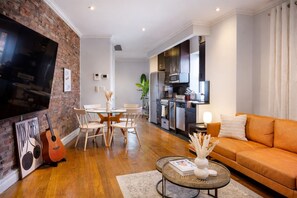 Un salon moderne avec un mur en briques, un téléviseur à écran plat, un coin repas doté d’une table en verre et une cuisine avec des meubles sombres et des appareils en acier inoxydable.