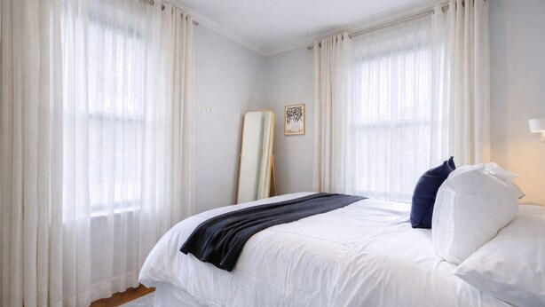 Una habitació d'hotel amb un llit, cortines blanques, una taula de nit i una làmpada penjada a la paret.