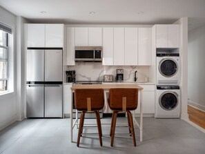 A modern kitchen with white cabinets, a stainless steel refrigerator, a microwave, a coffee maker, and a stacked washer and dryer. There are two wooden stools at a small kitchen island. - Renovated Luxury Three Bedroom Greenpoint Condo! (Brooklyn)