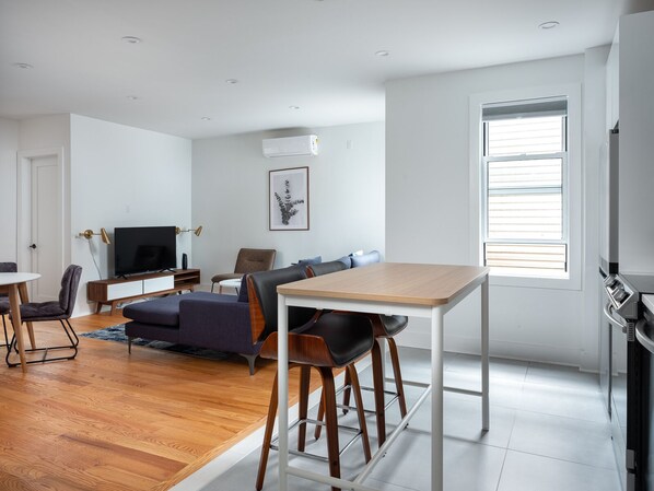 A modern living room with a wooden floor, a dining area with a table and chairs, a sofa, a TV, and a window with blinds. - Renovated Luxury Three Bedroom Greenpoint Condo! (Brooklyn)