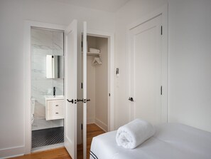 A modern bedroom with a neatly made bed, a bathroom with a marble vanity, and a hallway leading to a closet. - Renovated Luxury Three Bedroom Greenpoint Condo! (Brooklyn)