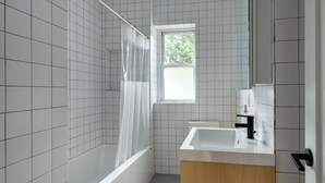 Un baño con paredes de azulejos blancos, una bañera, una ducha con cortina transparente, una ventana y un lavabo con espejo.