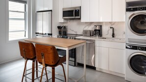 Cocina moderna con armarios blancos, microondas de acero inoxidable, cafetera y lavadora. Hay una mesa de madera con dos sillas.