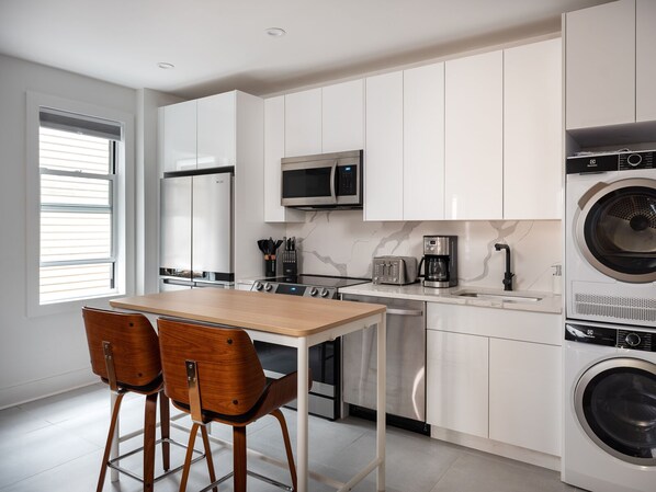 A modern kitchen with white cabinets, a stainless steel microwave, a coffee maker, and a washing machine. There is a wooden table with two chairs. - Renovated Luxury Three Bedroom Greenpoint Condo! (Brooklyn)